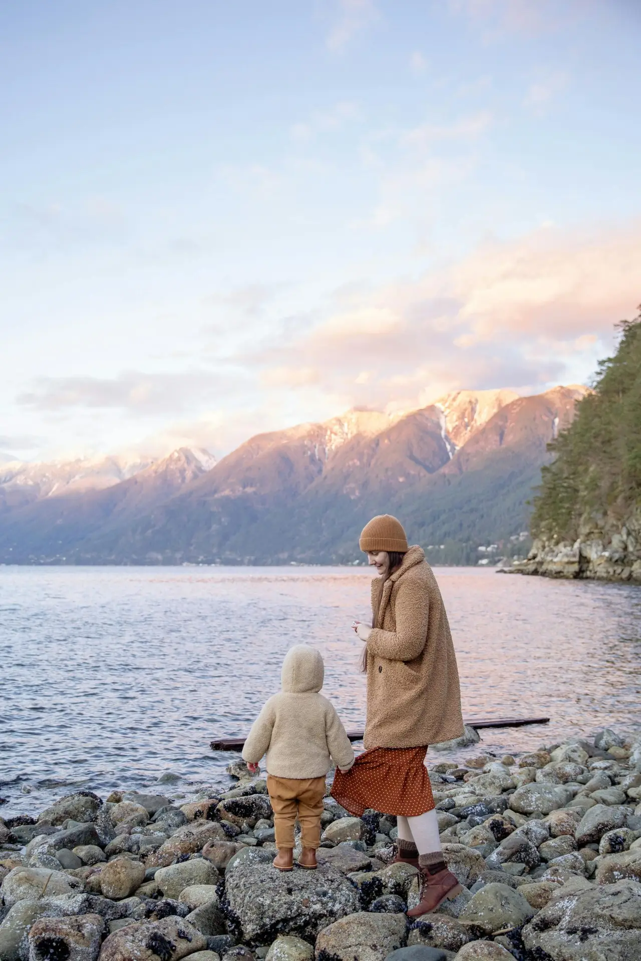 Back view unrecognizable preschool boy in warm outerwear holding skirt of mother in warm clothes and hat while traveling on calm lake in autumn